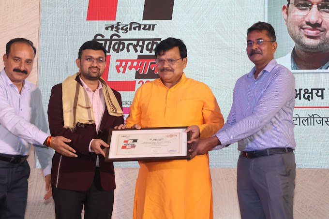 Four men are standing on a stage, three of whom are wearing shirts and one in an orange kurta. The man in the orange kurta is presenting a framed certificate or award to a man in a dark suit with a stole, while another man in a striped shirt stands to the left and a man in a blue shirt stands to the right. A large banner with Hindi text and a person's photo is in the background.
