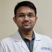 A close-up headshot of a man with short dark hair and glasses, wearing a white lab coat over a dark collared shirt, smiling subtly at the camera.