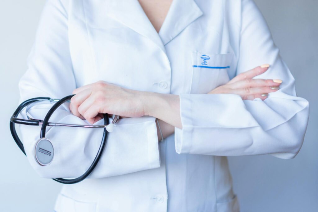 A close-up of a medical professional in a white lab coat with a blue medical caduceus logo on the pocket, holding a stethoscope with folded arms.