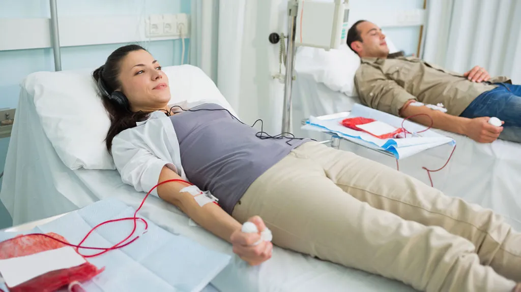Two people are lying on beds in a medical setting, each with an IV line in their arm, appearing to be donating blood.
