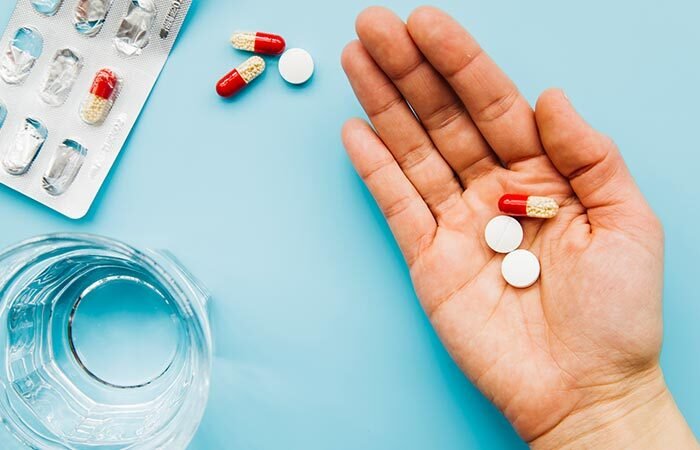 A hand holds two white pills and a red and white capsule, with more pills, a blister pack, and a glass of water on a light blue surface.