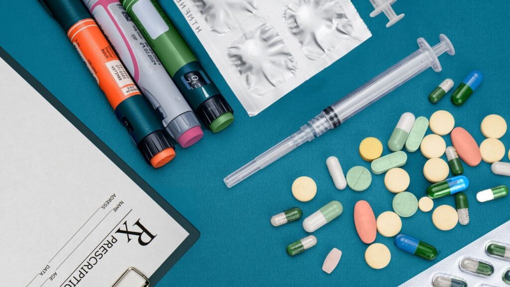 A flat lay of various medications, including pre-filled injector pens, blister packs, a syringe, and scattered pills and capsules, alongside a prescription pad.