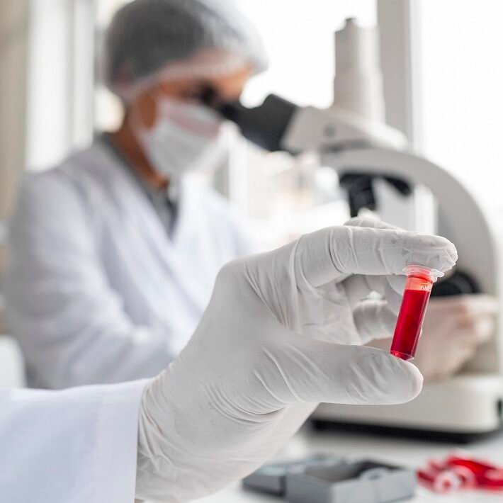 Scientist in protective gloves holding a small vial filled with red liquid, with a microscope and a blurred lab technician in the background.